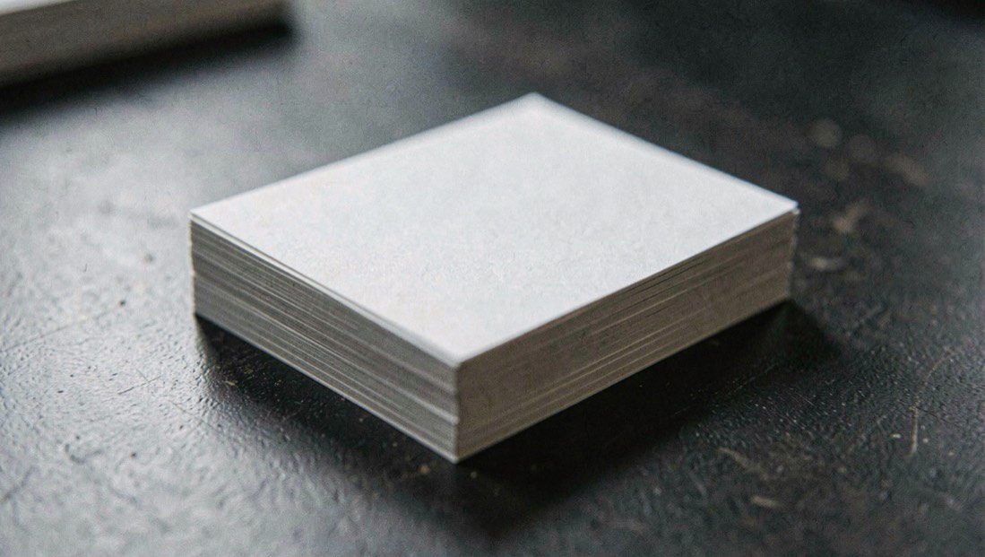 Une pile de cartes imprimées vierges posée sur un plan de travail sombre, angles vifs bien nets, lumière rasante qui détache l’épaisseur, macro.