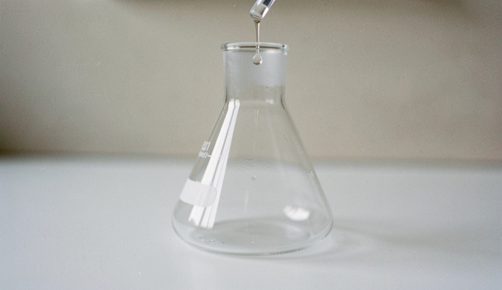 Un entonnoir de laboratoire en verre posé sur une paillasse blanche, goutte suspendue au col, fond neutre, lumière douce, macro.