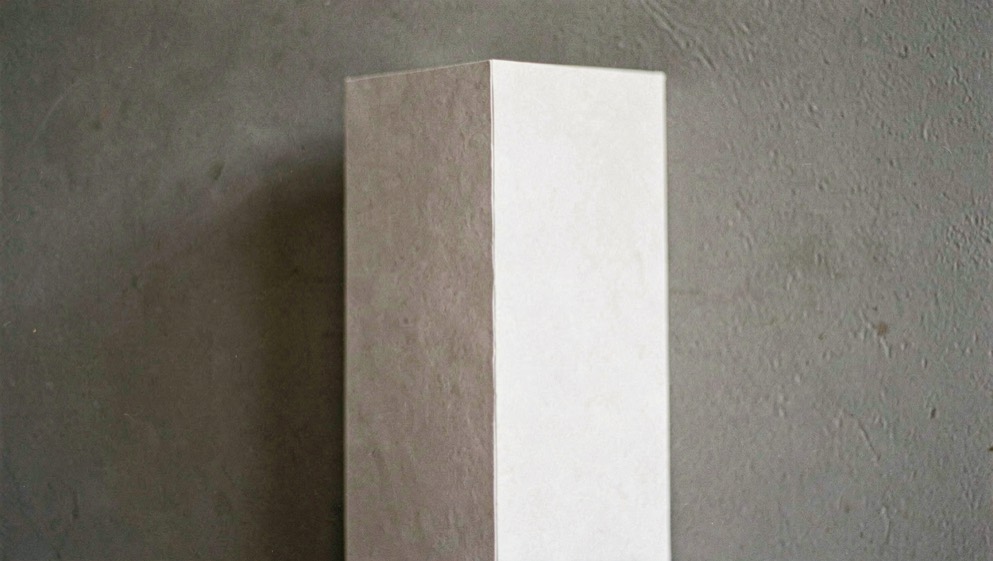 Une feuille blanche pliée en deux tenue verticalement contre un fond gris uni, pli net au centre, lumière latérale douce, plan serré.