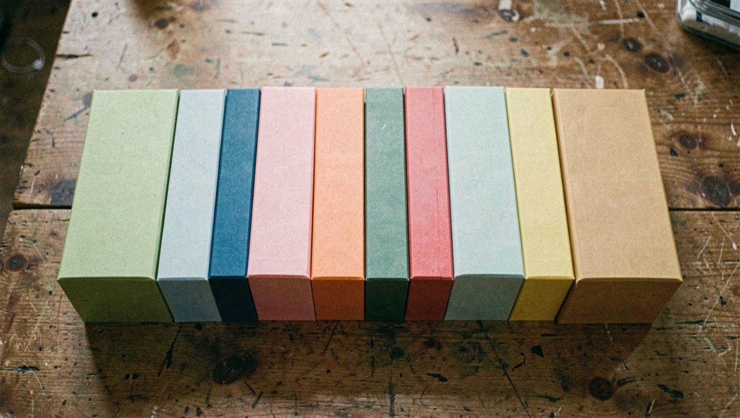 Des cartons épais de couleurs unies, sans impression, alignés côte à côte sur une table en bois, vue de dessus, ombres douces.